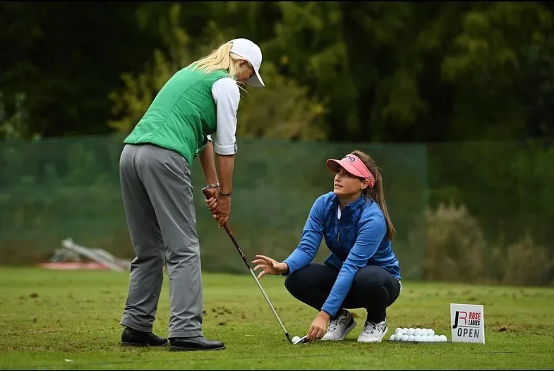 Jae Bowers crouching to adjust a player's grip on the practice ground at the Rose Ladies Open